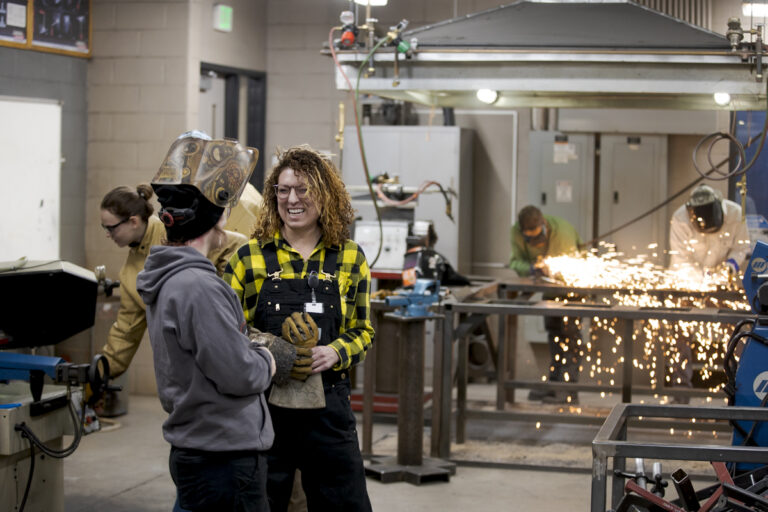 Welding Students Converse and Laugh with Each Other as Other Students Weld in the Background with Sparks Flying in Denver, CO