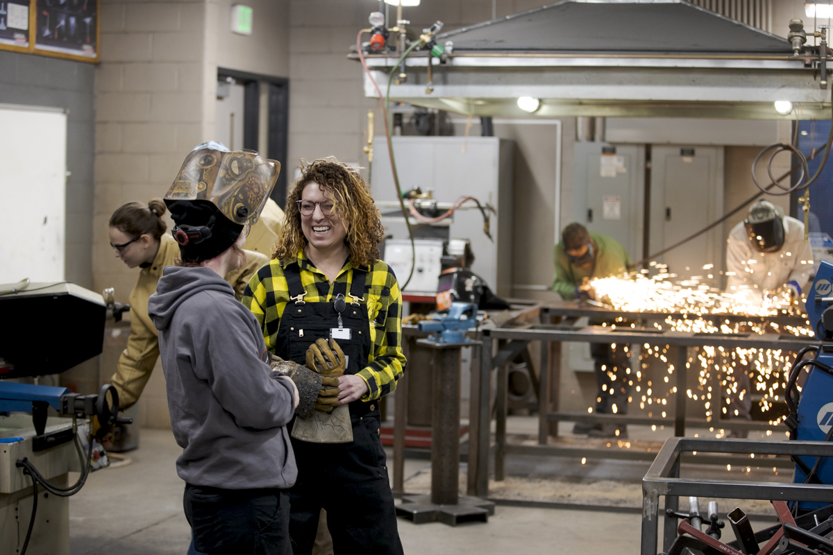 Welding Students Converse and Laugh with Each Other as Other Students Weld in the Background with Sparks Flying in Denver, CO