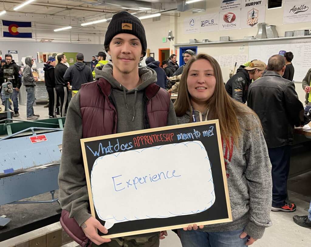 Apprenticeship students smiling while holding a board asking "what does apprenticeship mean to me?" Students' answer is "experience"