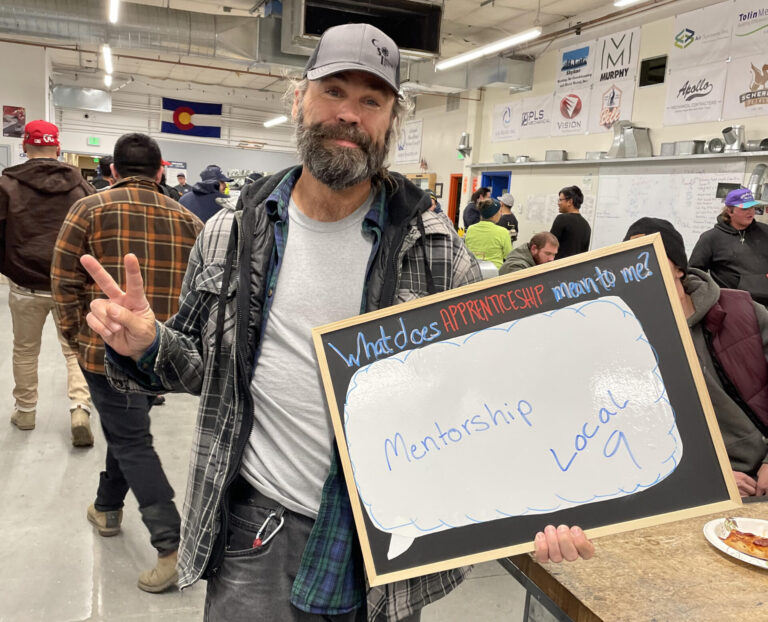 Apprenticeship student smiling while holding a board asking "what does apprenticeship mean to me?" Student's answer is "mentorship"